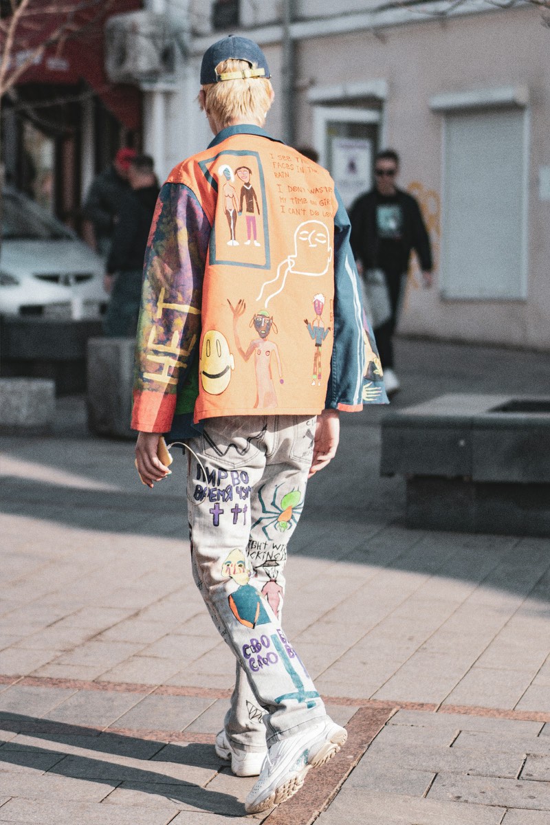 A person photographed from behind on a city pavement, wearing a backwards cap, a hand-painted orange patchwork jacket, painted light trousers and white trainers.