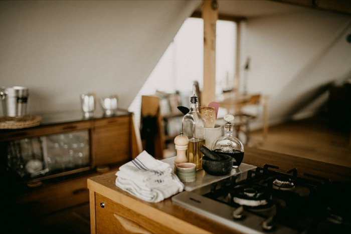 A home kitchen counter with a gas hob, olive oil bottle, pepper mill, stone mortar and pestle, and folded tea towels.